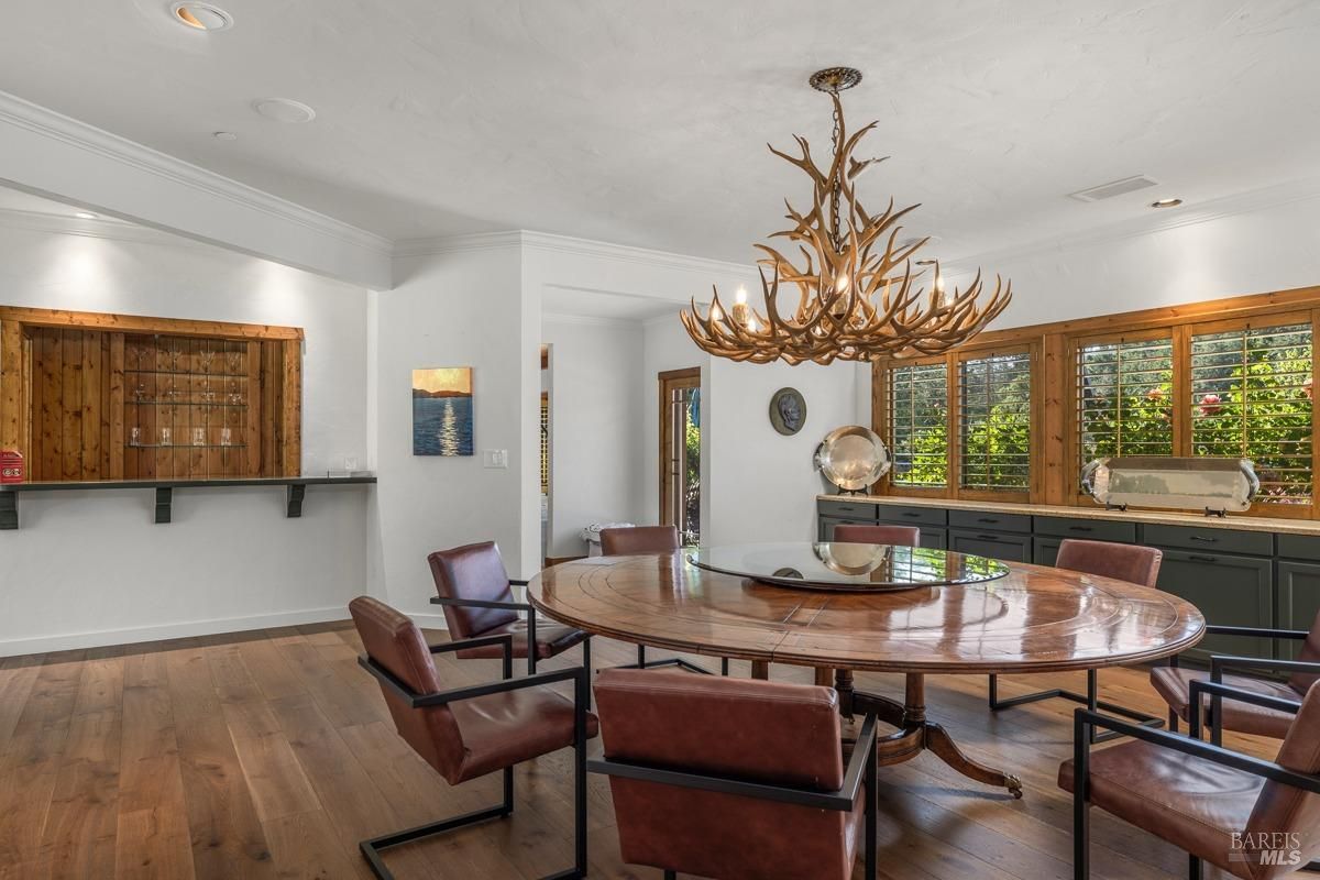 A spacious and elegant dining room with a large round table, leather chairs, and a stunning antler chandelier. The room features large windows with wooden shutters, providing plenty of natural light.