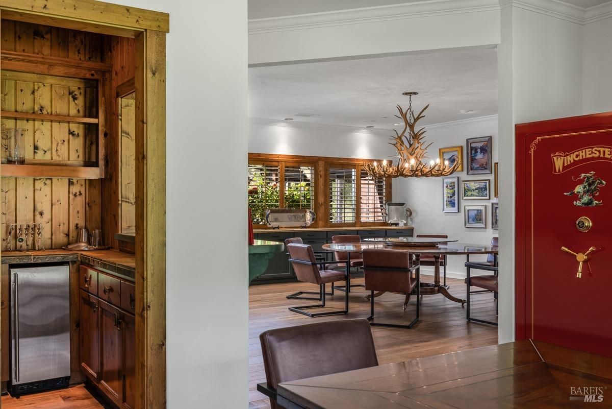 A stylish dining room with a large round table, leather chairs, and a unique antler chandelier. The room features a built-in bar area and a large safe, adding a touch of luxury and security.