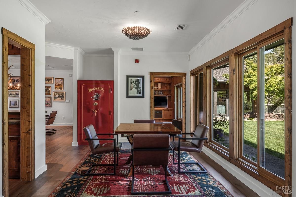 A cozy and inviting dining nook with a small table, leather chairs, and a colorful rug. The room features large windows with beautiful views of the outdoors and a large, red safe.