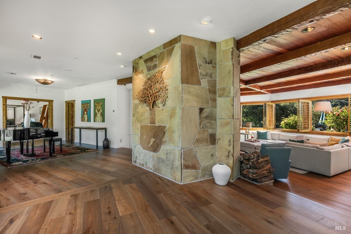  A transitional living space with wood flooring and a prominent stone wall with an artistic tree design. The open plan connects this area to a seating zone with ample natural light and wood-beamed ceilings.