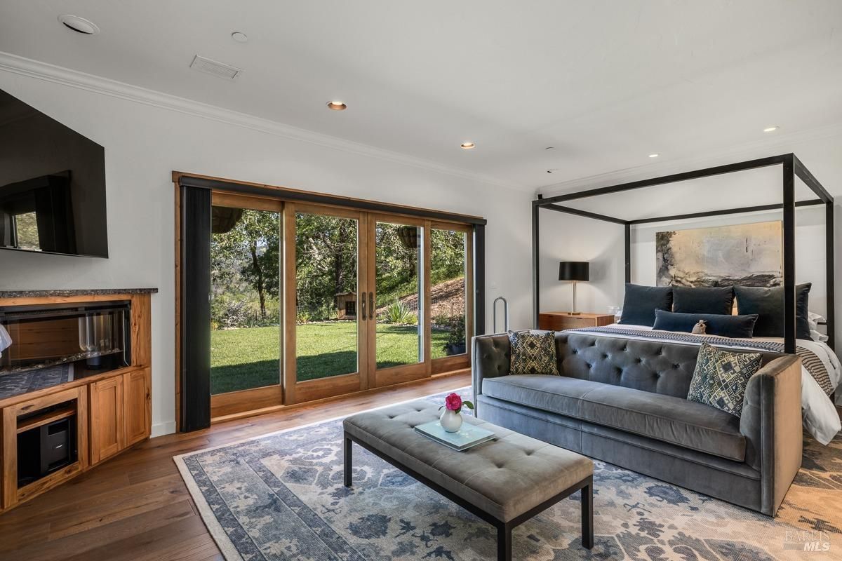 A spacious and luxurious bedroom with a large bed, a cozy sitting area with a sofa and coffee table, and a fireplace. Large sliding glass doors open onto a beautiful outdoor view. The room is decorated in a modern style with a touch of rustic charm.