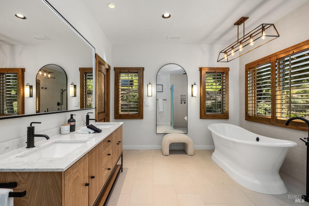 A spacious bathroom with a modern freestanding bathtub and double vanity with wood cabinetry. The arched mirror and wall-mounted lights add elegance, while the plantation shutters frame the windows beautifully. 