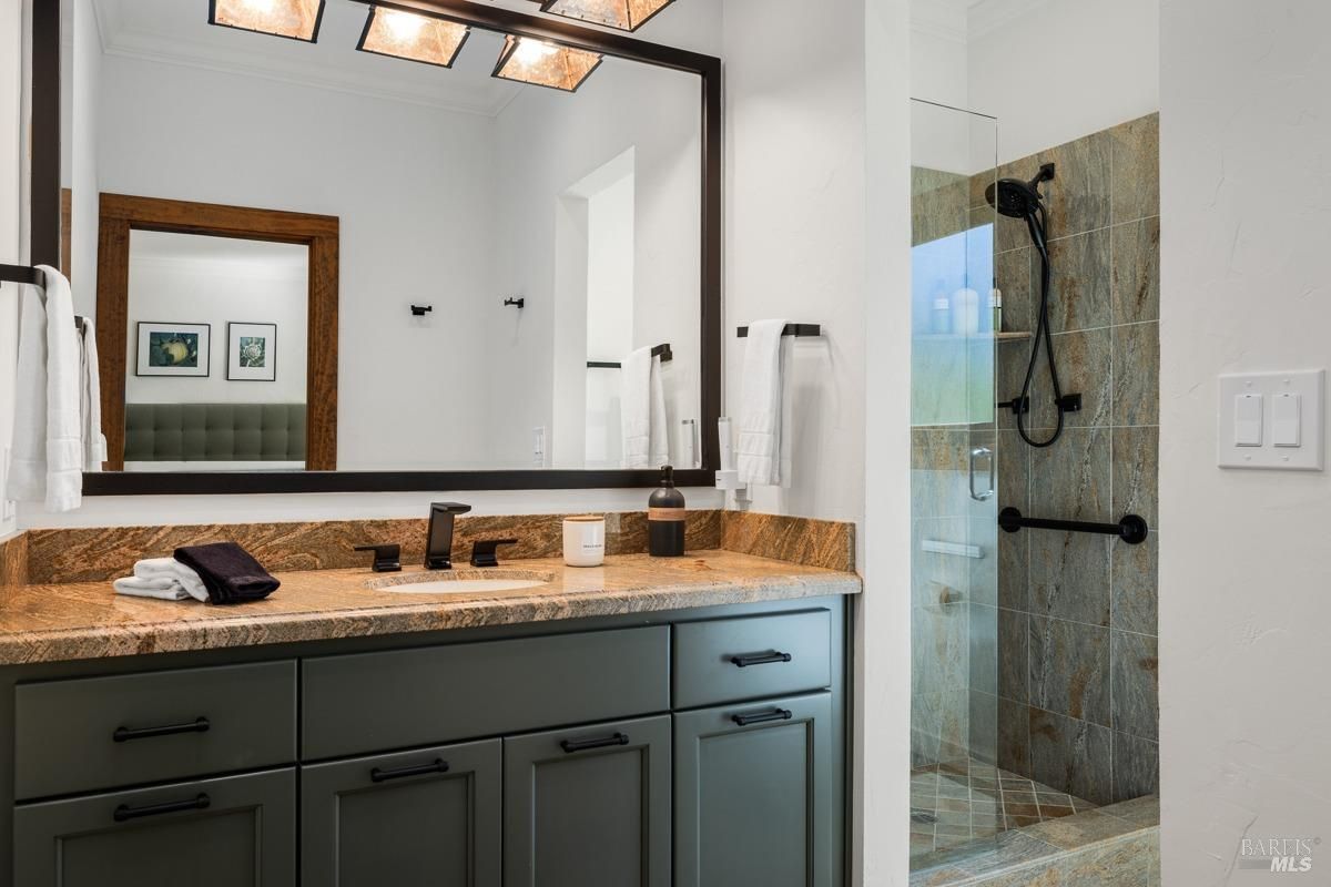 This bathroom has a warm, earthy aesthetic with a granite countertop, green cabinetry, and a walk-in shower with natural stone tiles. Black fixtures and a large framed mirror complete the look.