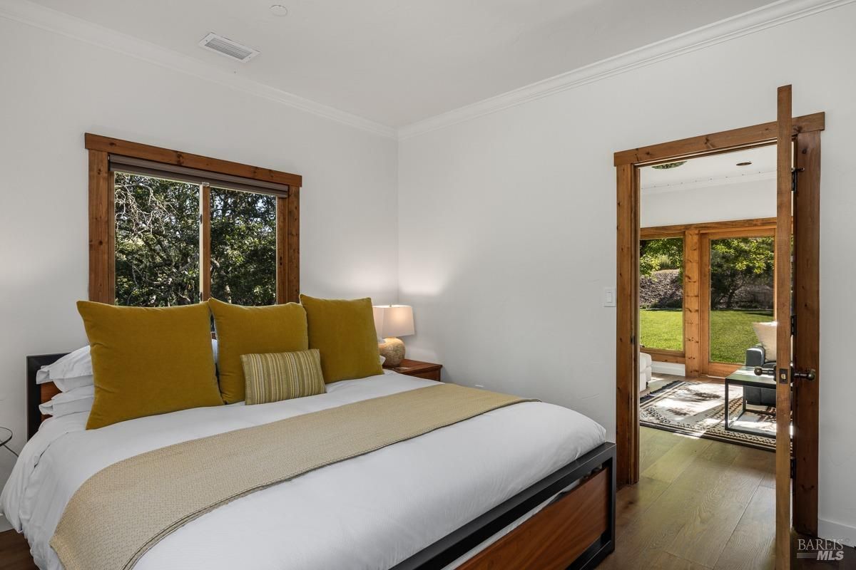 This cozy bedroom features a simple yet elegant design with mustard-yellow pillows as the focal point. The room has large wooden-framed windows and an adjoining space that opens to a bright, naturally lit area. The neutral color palette complements the natural surroundings.