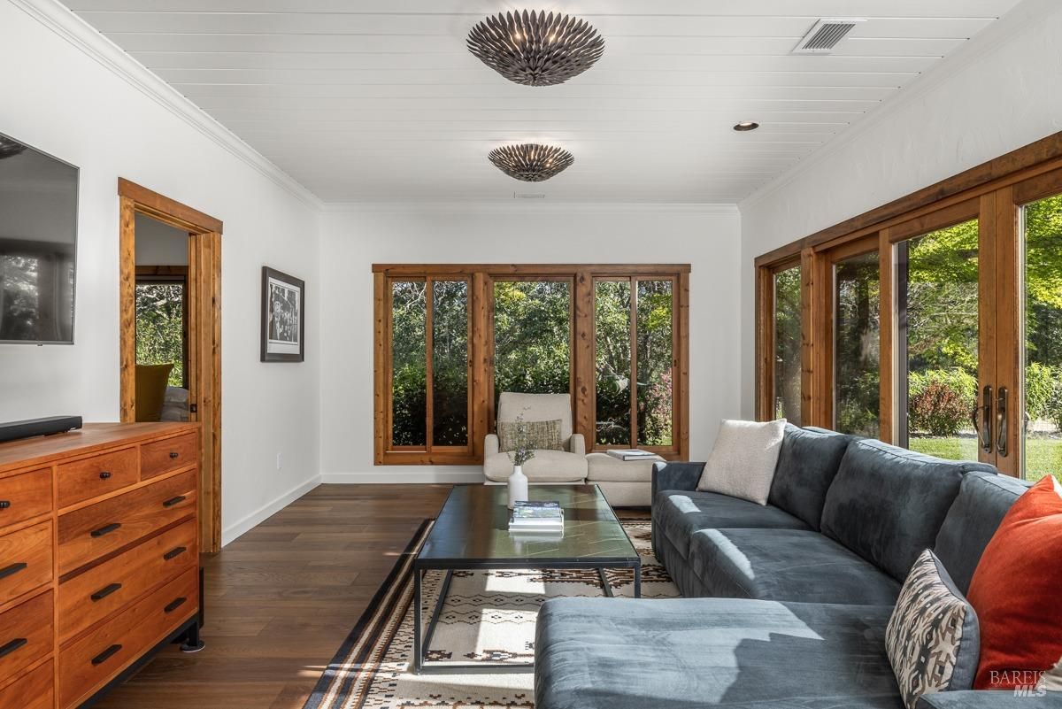 A cozy living room with a comfortable couch, a coffee table, and a pair of armchairs. The room features large windows that offer plenty of natural light and views of the outdoors. The wooden ceiling adds a touch of warmth and character to the space.