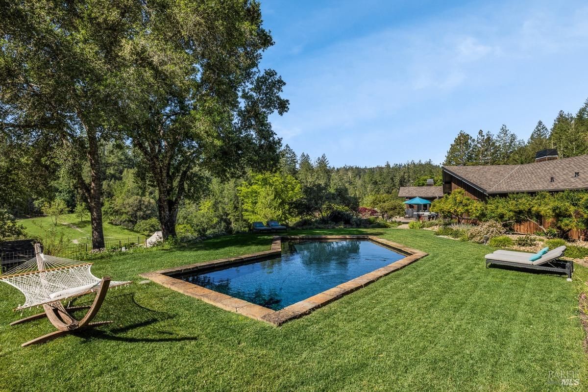  A lush backyard featuring a rectangular stone-edged pool surrounded by green lawns and mature trees. A hammock and lounge chairs are thoughtfully placed for relaxation, creating an inviting and tranquil outdoor space.