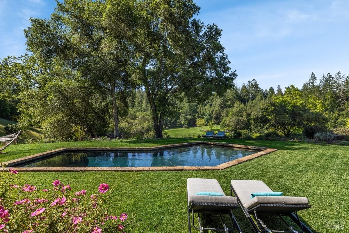 Pool area, showing blooming pink flowers in the foreground and expansive greenery in the background. The pool, flanked by lounge chairs, is set in a serene natural setting.