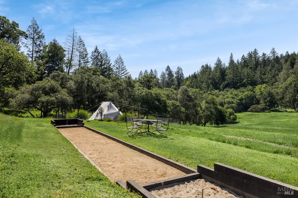 A bocce ball court sits nestled amidst a lush green lawn, surrounded by towering trees and a picturesque landscape. A small tent and a picnic table add to the outdoor ambiance.