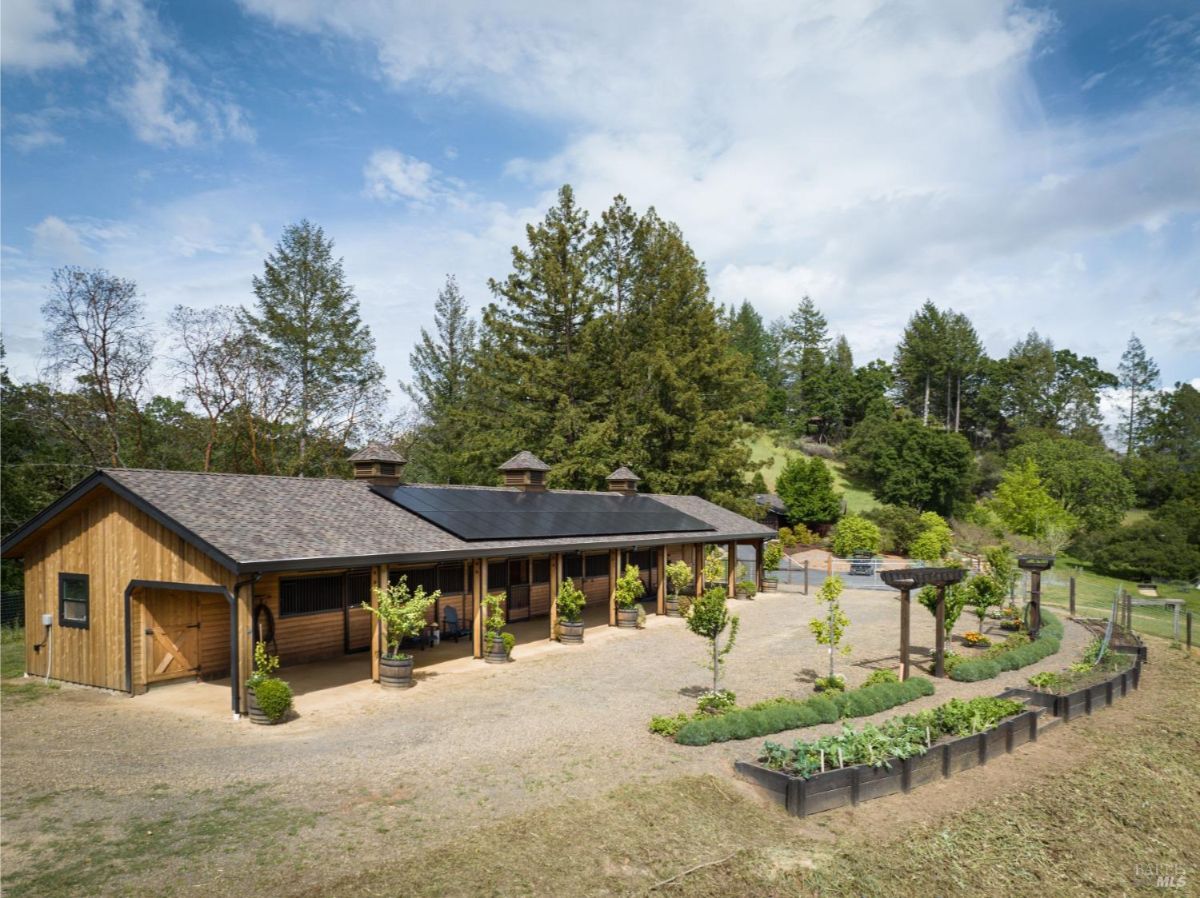 A broad view of a stable building featuring solar panels on the roof. The stable is accompanied by neatly arranged planter boxes filled with various plants or crops. The surrounding area includes trees and open land, indicating a sustainable and eco-friendly ranch.