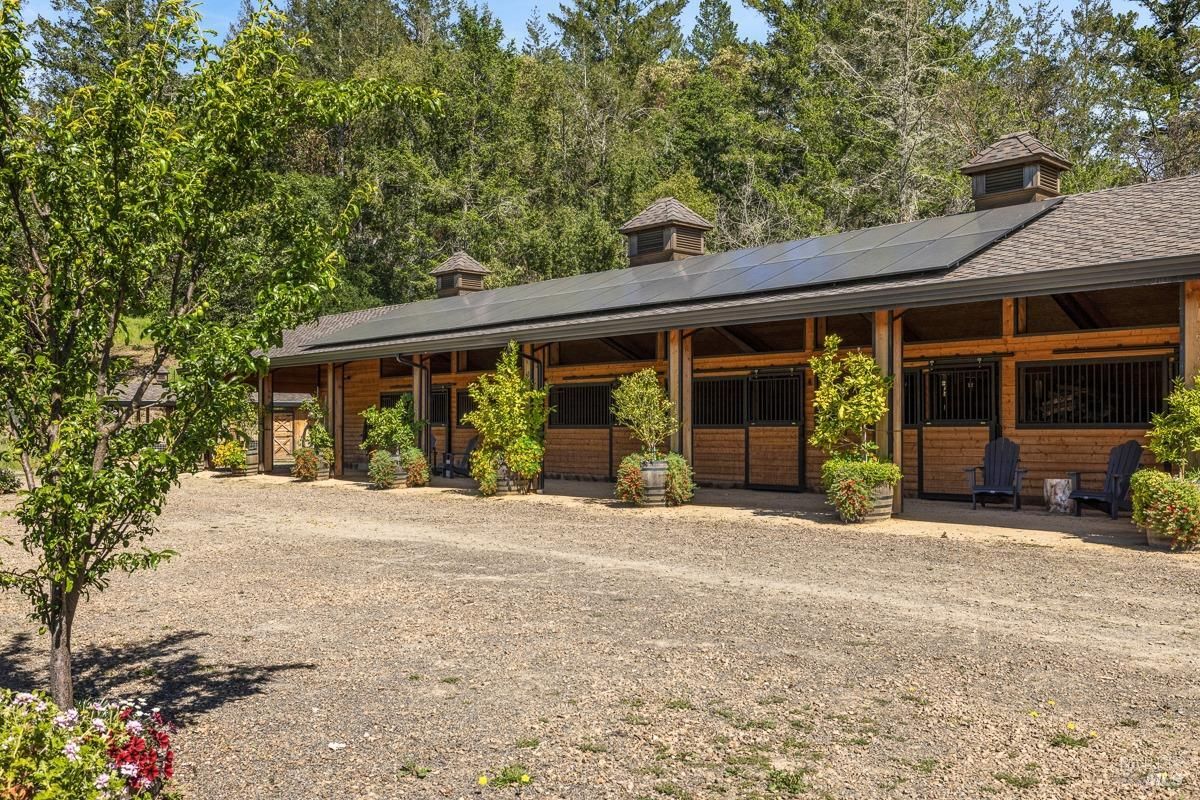 A horse stable from the exterior, showcasing a long structure with solar panels on the roof and wooden paneling. The surrounding area includes trees and open land, combining sustainability with rural aesthetics.