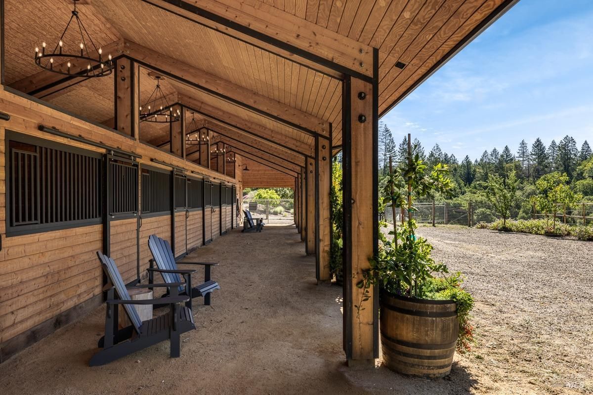 A horse stable with a wood and metal structure, featuring a row of stalls and Adirondack chairs. The design blends rustic charm with modern utility, set against a backdrop of open landscapes.