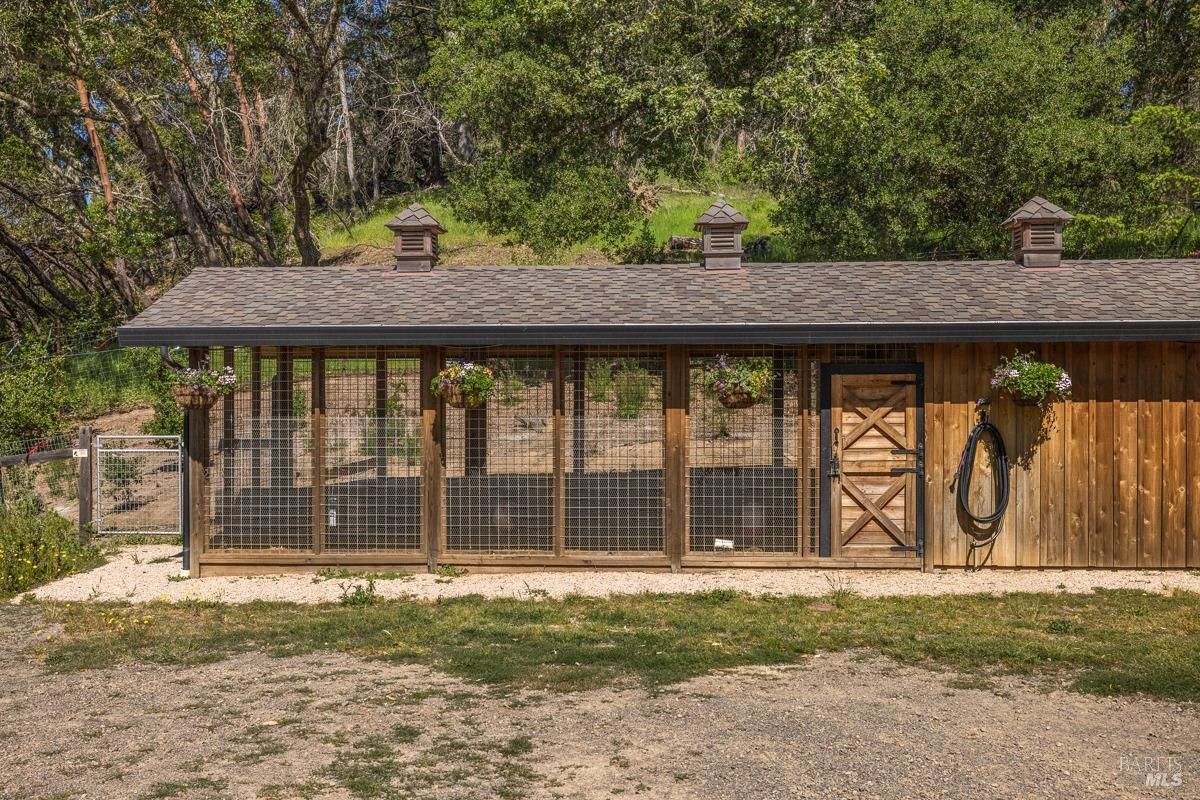 The exterior of the chicken coop, a neatly constructed wooden structure with hanging flower baskets. It blends functionality with aesthetic charm, surrounded by greenery.