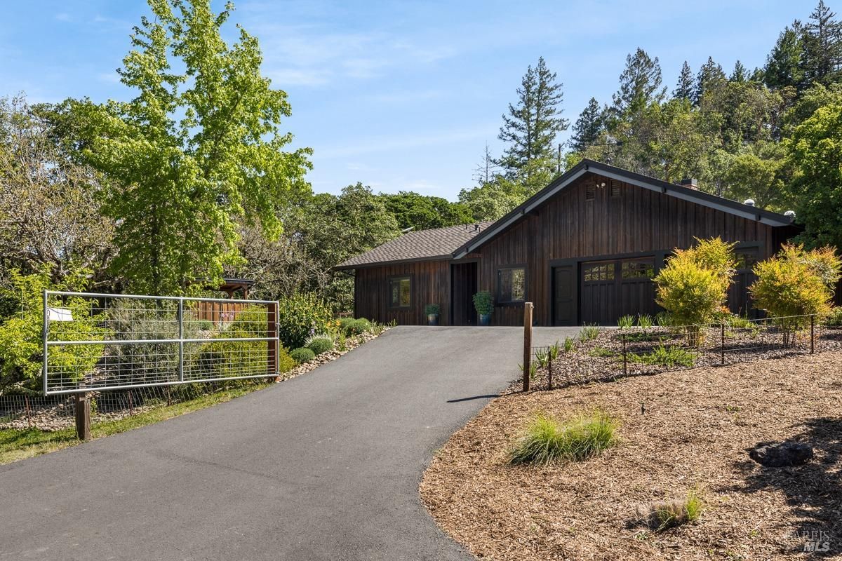 A wooden house or ranch-style building with a modern, minimalist design. A paved driveway leads to the building, and there are decorative plants and shrubs around the area. The setting appears to be a private residence or guesthouse on a rural property.