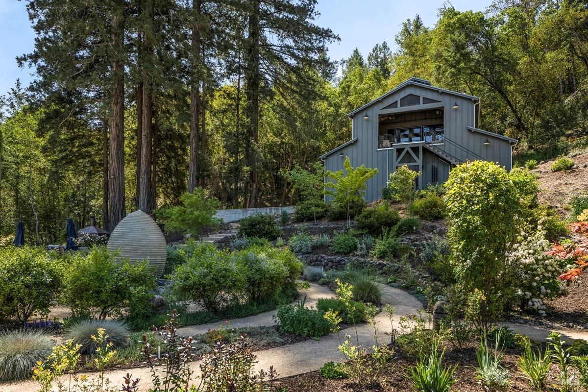 A picturesque garden path winds through a lush, green landscape, leading to a charming barn nestled amidst tall trees. The garden features terraced beds, stone walls, and a variety of colorful flowers.