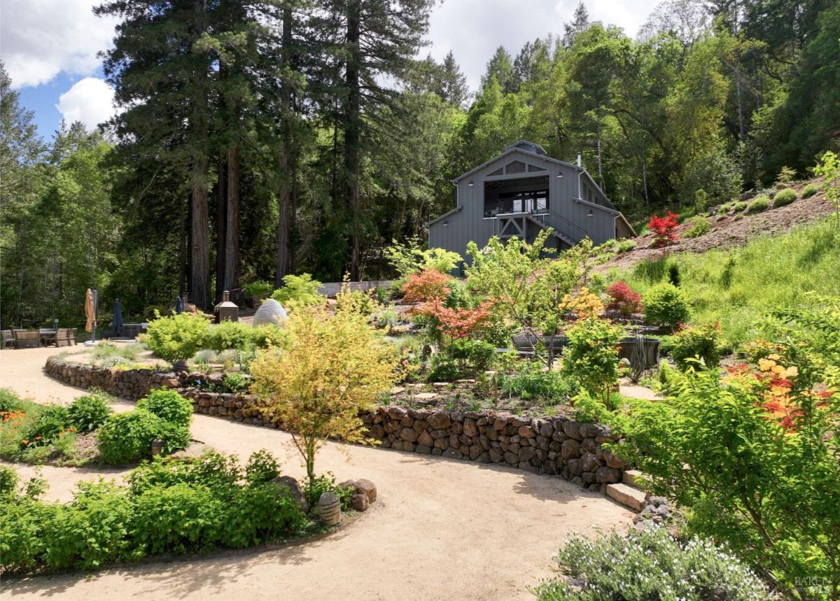 A picturesque garden path winds through a lush, green landscape, leading to a charming barn nestled amidst tall trees. The garden features terraced beds, stone walls, and a variety of colorful flowers.