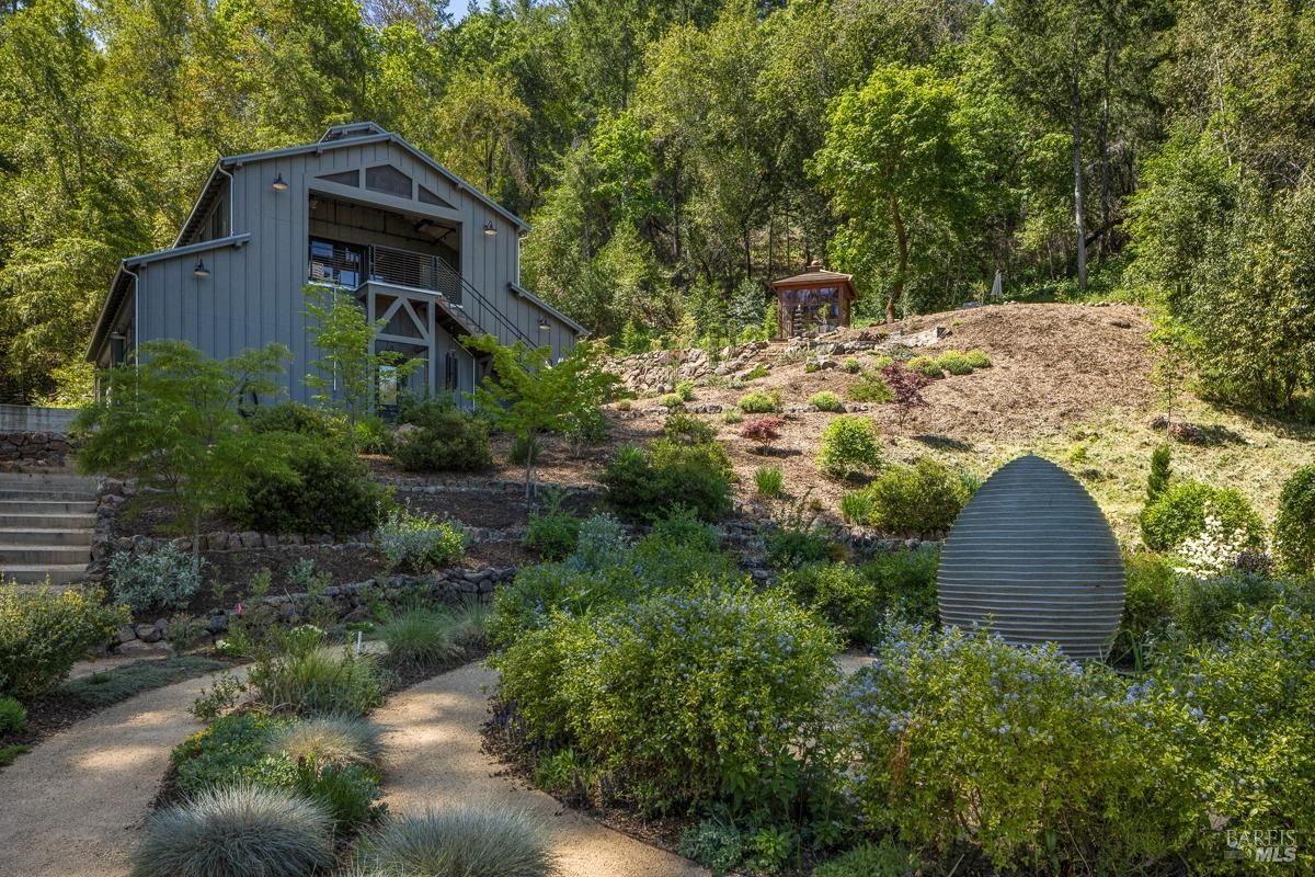 A large barn-like structure with a contemporary design, situated on a hillside. Surrounding the building are landscaped gardens and pathways, along with a modern artistic installation in the foreground. The setting blends rustic architecture with modern landscaping elements.