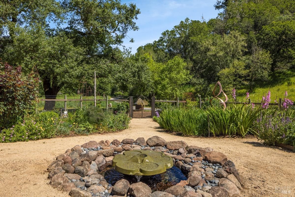 A small fountain with a floral-shaped centerpiece, surrounded by smooth stones and lush greenery. In the background, a wooden gate leads to a garden area, enhancing the serene and natural ambiance.