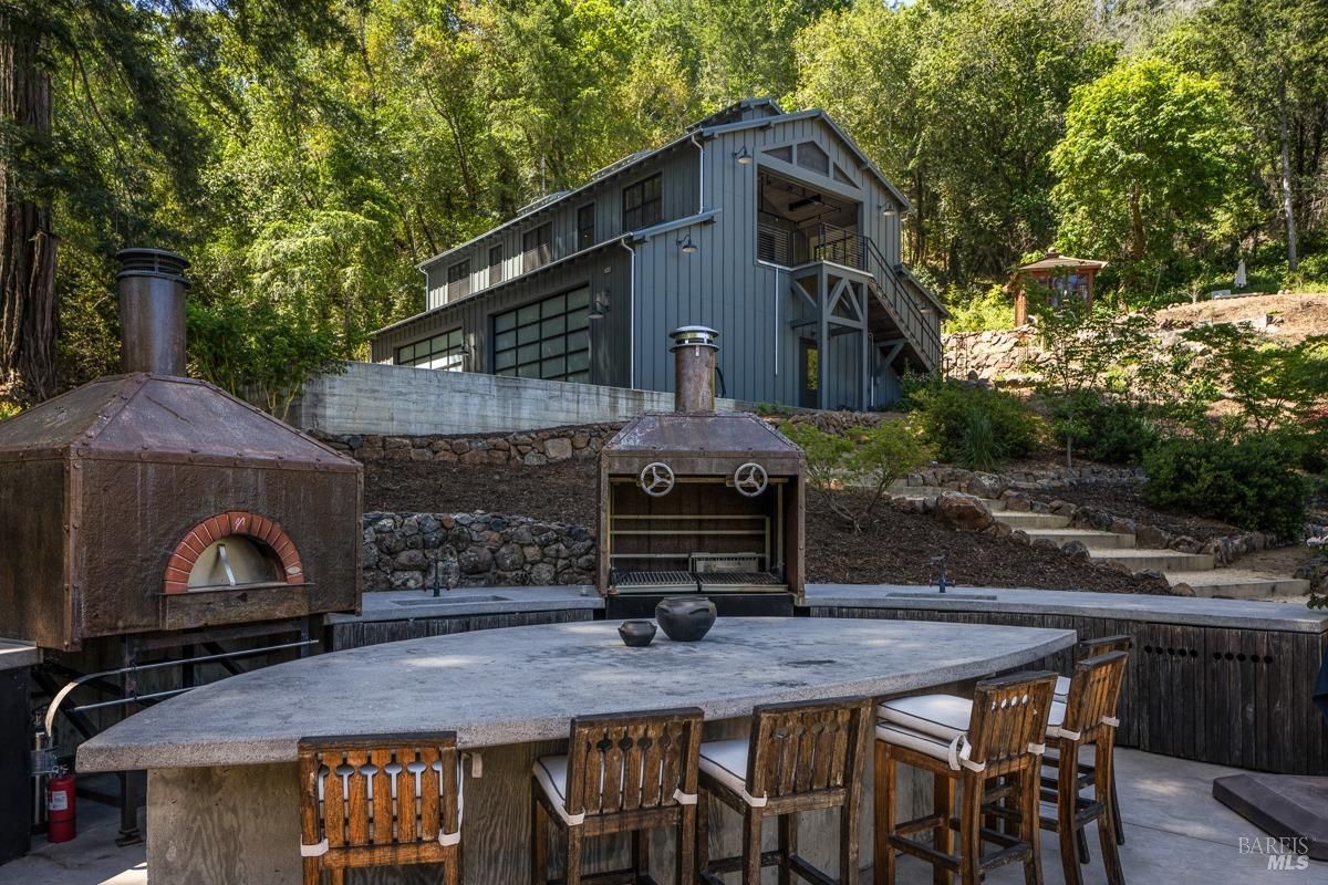 : A rustic outdoor kitchen with a wood-fired oven, grill, and a curved bar-style dining table with wooden chairs. The backdrop features a modern barn-like structure surrounded by lush greenery.