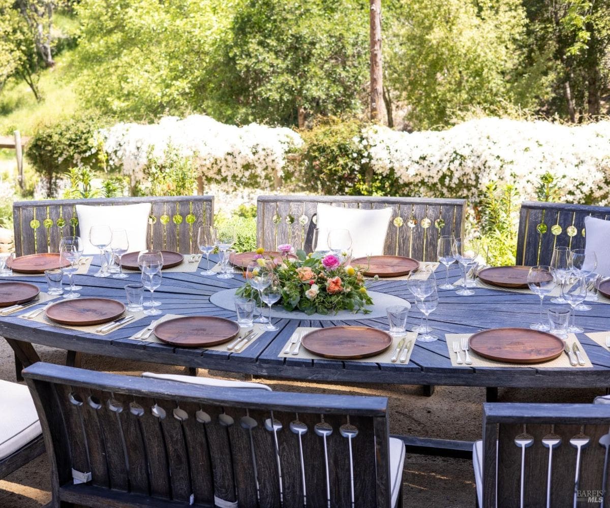 A charming outdoor dining table set for a gathering, with wooden chairs and plates arranged around a floral centerpiece. The setting is surrounded by blooming flowers and greenery, creating a festive and picturesque ambiance.