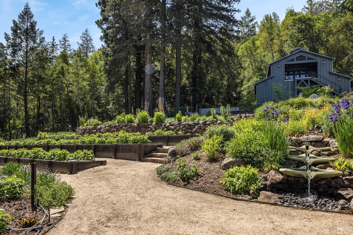 Garden path winds through a lush, green landscape, leading to a charming barn nestled amidst tall trees. The garden features terraced beds, stone walls, and a cascading water feature.