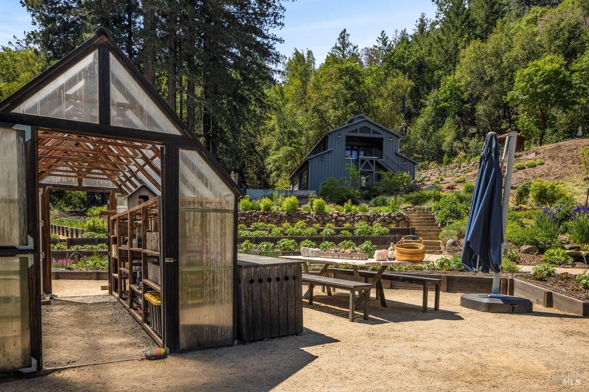  A greenhouse structure with transparent panels, surrounded by a well-maintained garden featuring vibrant plants and flowers. A picnic table and umbrellas are visible, suggesting an outdoor gathering or gardening area.