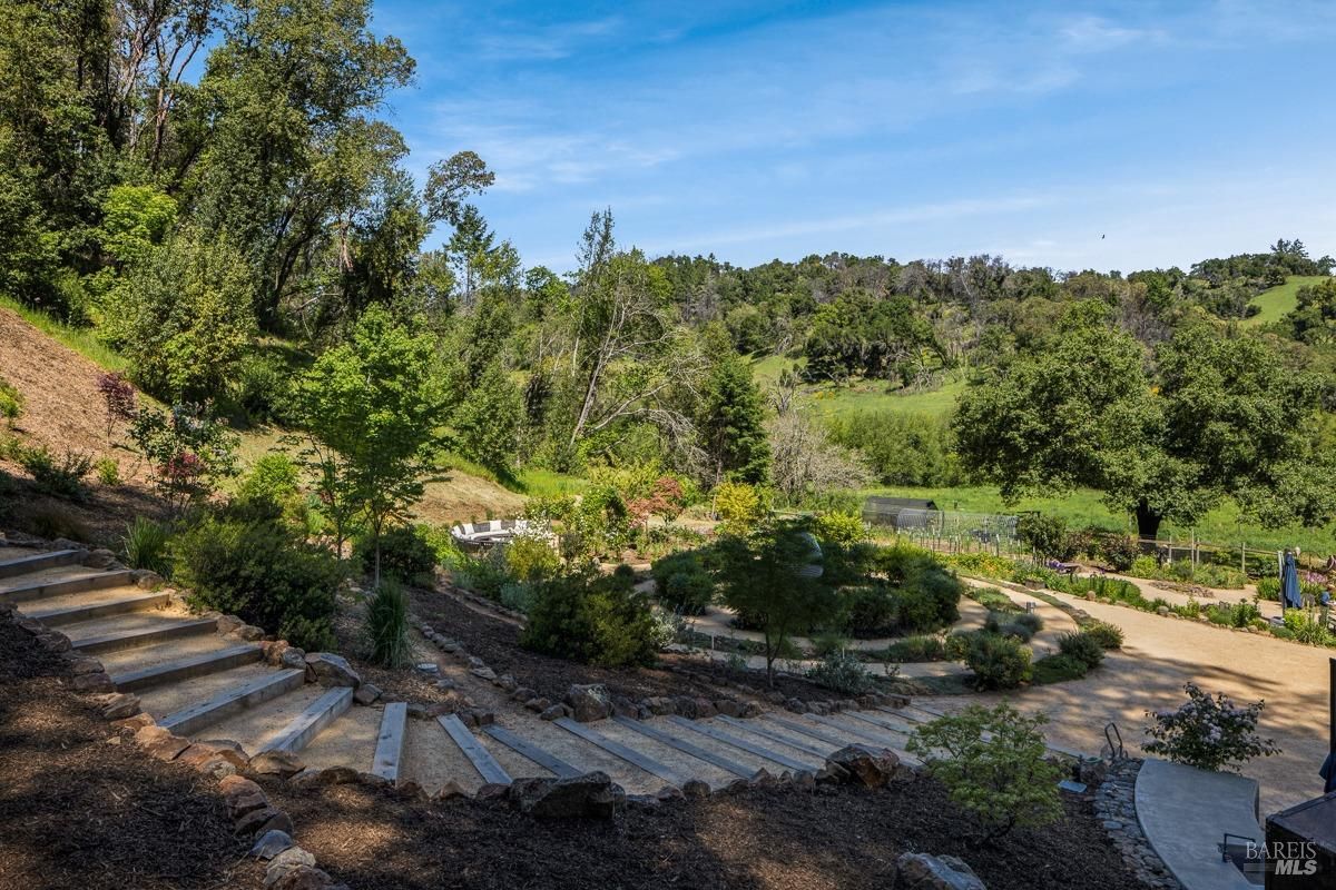 A terraced garden with stone steps leading downward. The garden features various plants and shrubs arranged in a landscaped design, with a backdrop of trees and open fields, creating a calming and aesthetic space.