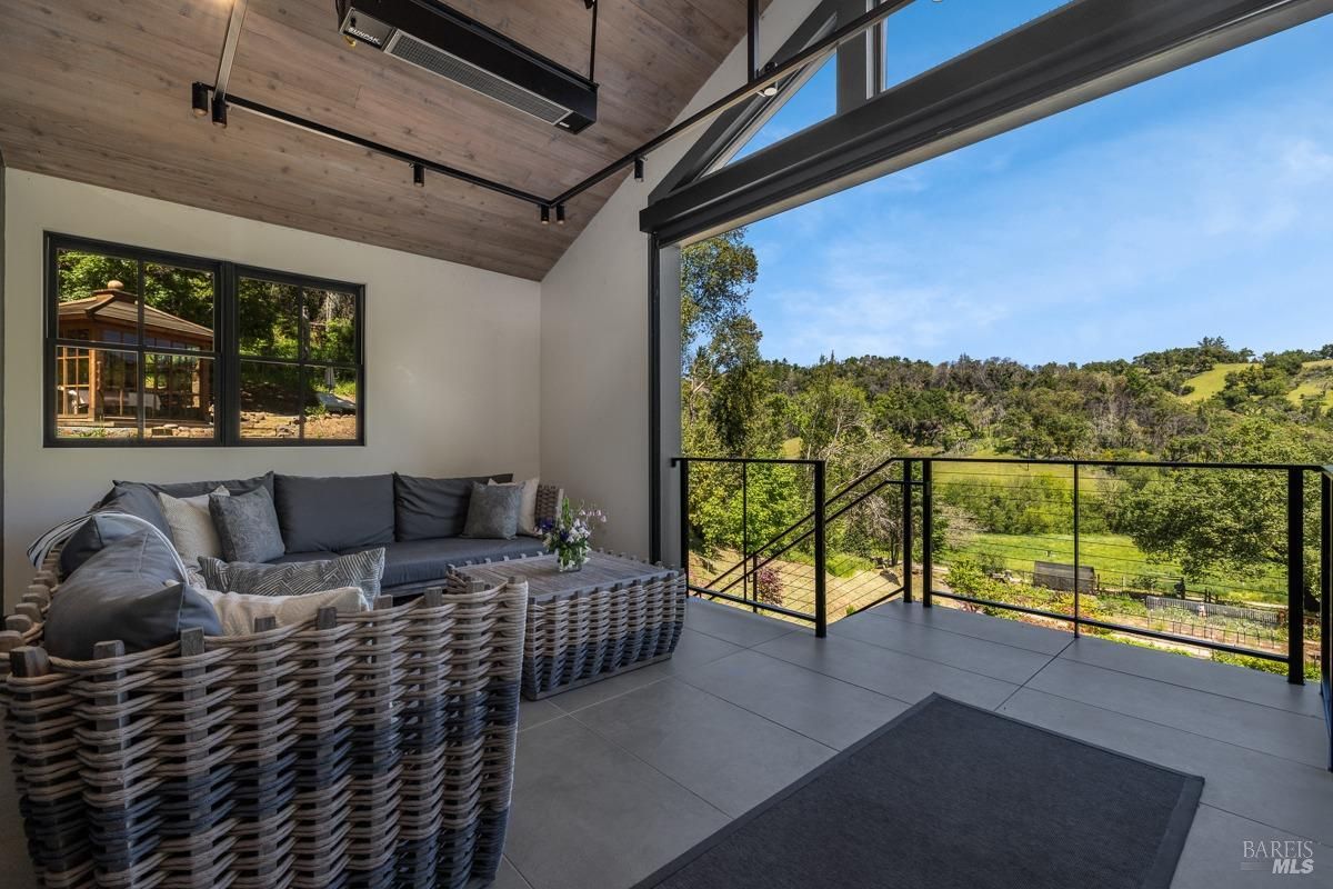  A cozy seating area on a balcony or patio, with gray wicker furniture and soft cushions. This space is perfect for relaxing while enjoying the scenic views of lush trees and hills. The design incorporates a modern wooden ceiling and ample natural light.