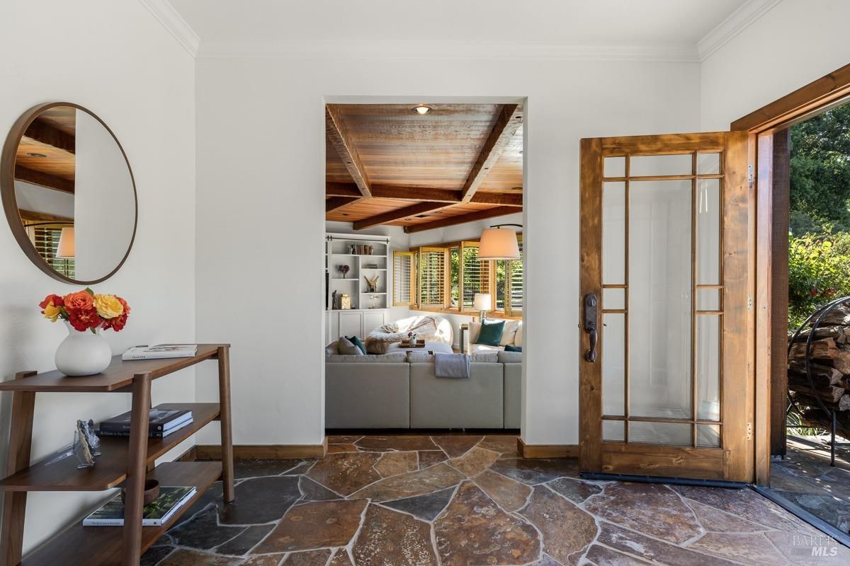 A bright and inviting entryway with a stone tile floor, a wooden console table, and a round mirror. The doorway leads into a cozy living room with a wood-beamed ceiling and large windows.