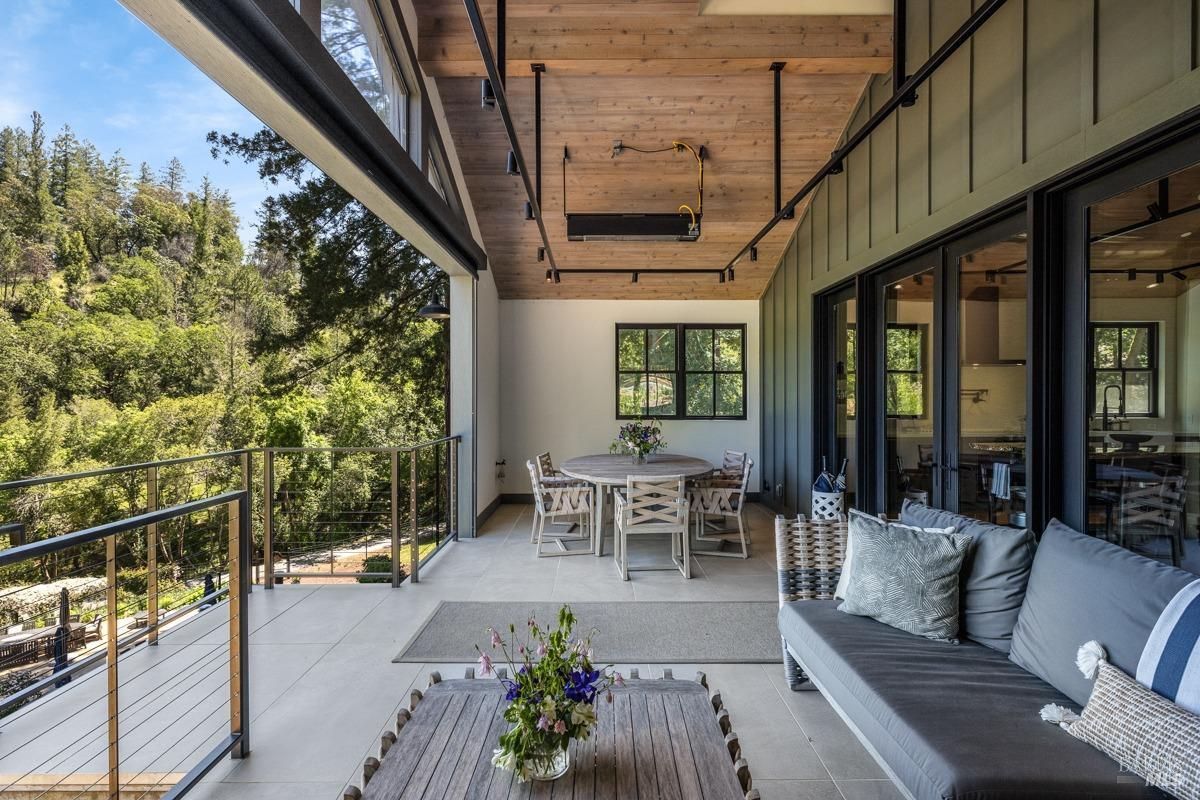  A spacious patio area featuring a round dining table with chairs and a comfortable seating area with cushioned wicker furniture. The patio is enclosed with glass railings, offering breathtaking views of the surrounding greenery. The wooden ceiling and modern lighting add warmth to the open-air design.