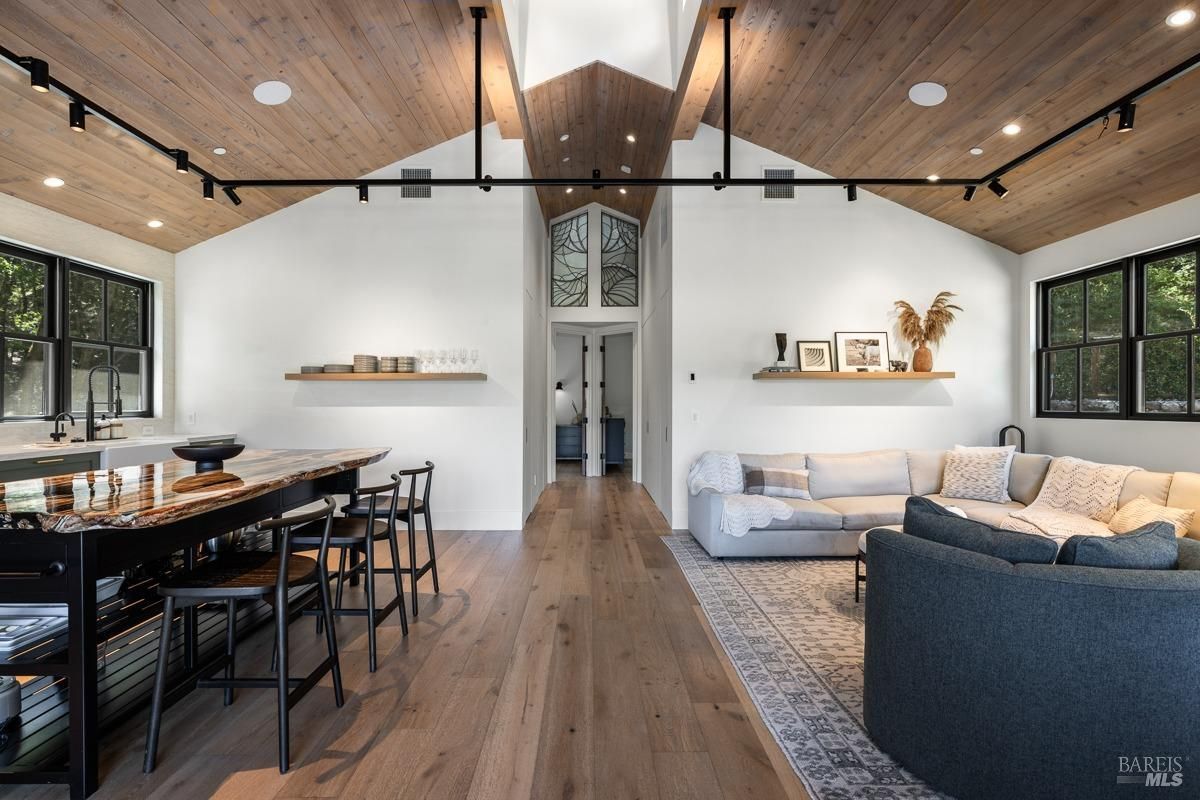 A modern and open-concept living space with a kitchen, dining area, and living room seamlessly flowing into each other. The room features a vaulted ceiling with exposed wooden beams, large windows, and a neutral color palette.