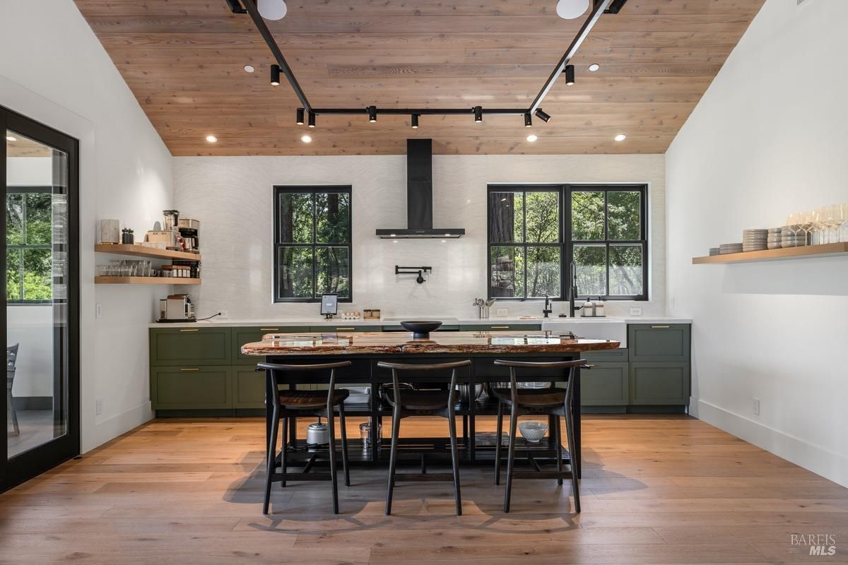 A modern and stylish kitchen with dark green cabinets, a large island with a wooden countertop, and black bar stools. The room features a vaulted ceiling with exposed wooden beams, large windows, and a sleek black range hood.