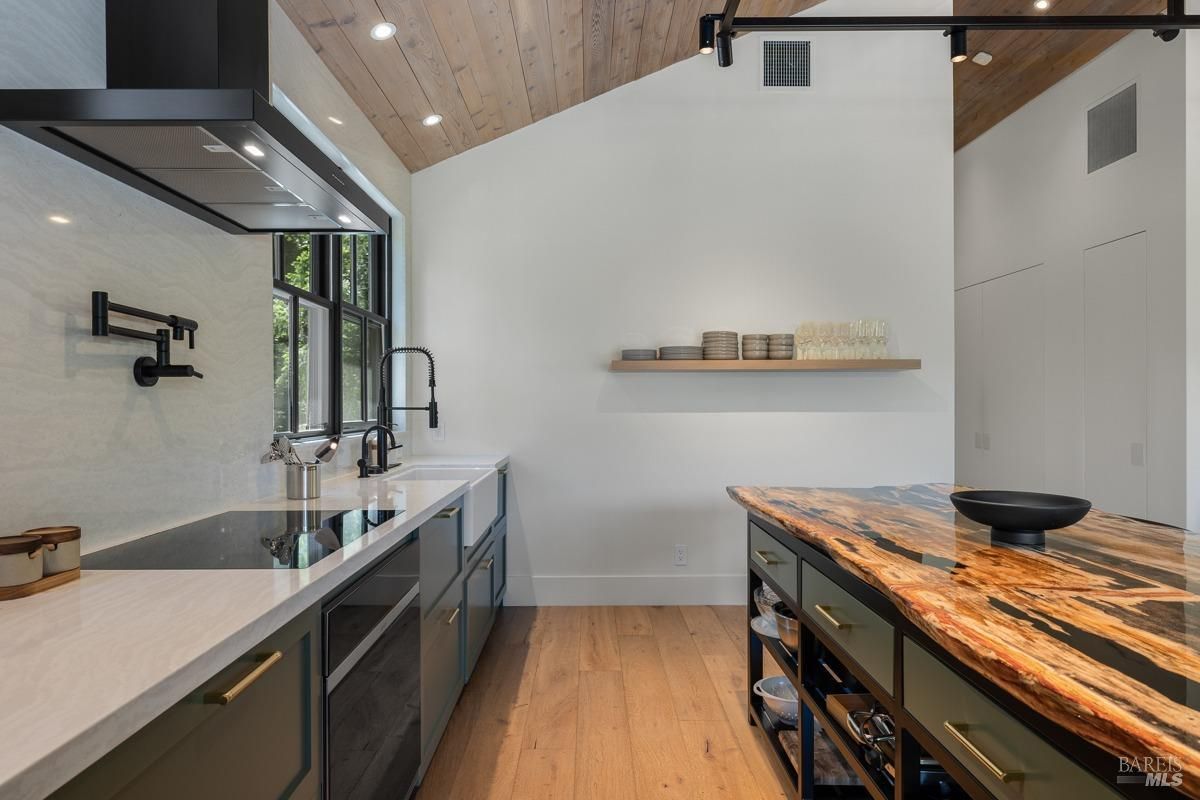 A sleek, minimalist kitchen featuring a mix of dark cabinetry, a marbled countertop, and a distinctive live-edge wood island countertop. The space is accentuated with black hardware and fixtures, large windows, and a vaulted ceiling with wood paneling for a contemporary yet natural look.