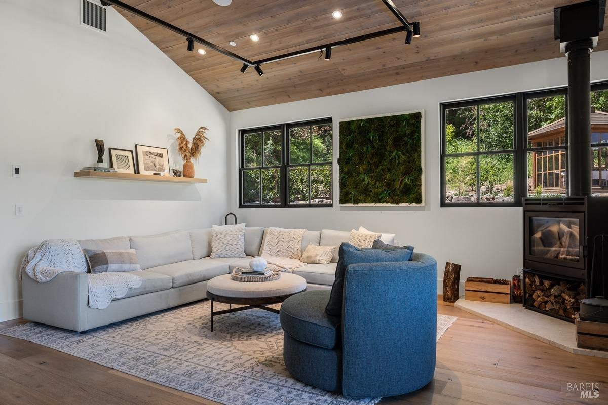 A cozy living room with a large sectional sofa, a blue armchair, and a coffee table. The room features a wood-burning fireplace, large windows with views of the outdoors, and a wood-beamed ceiling.