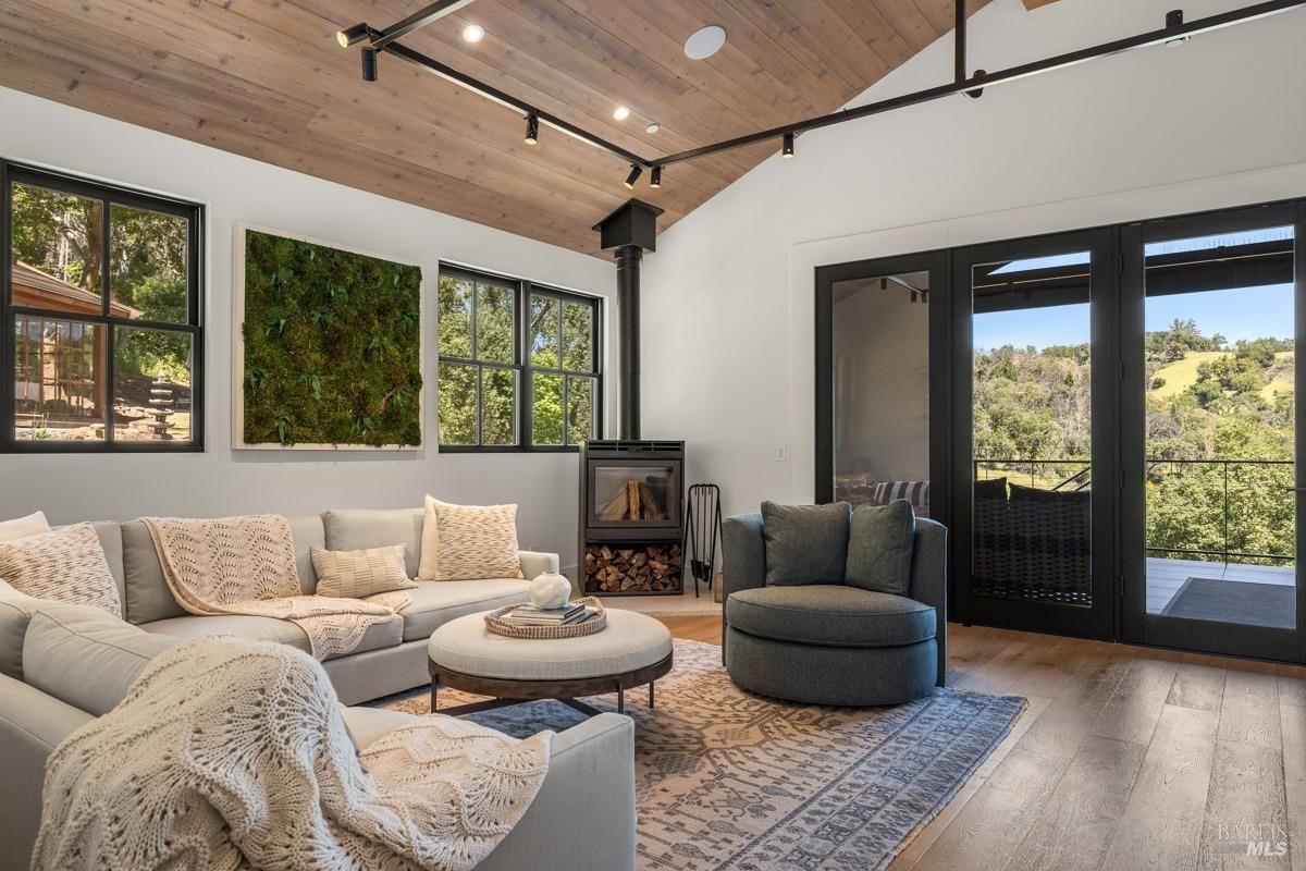 A spacious and inviting living room with a cozy sectional sofa, a comfortable armchair, and a fireplace. Large windows offer stunning views of the surrounding landscape, and sliding glass doors lead out to a deck or patio. The room features a wood-beamed ceiling and a modern design aesthetic.