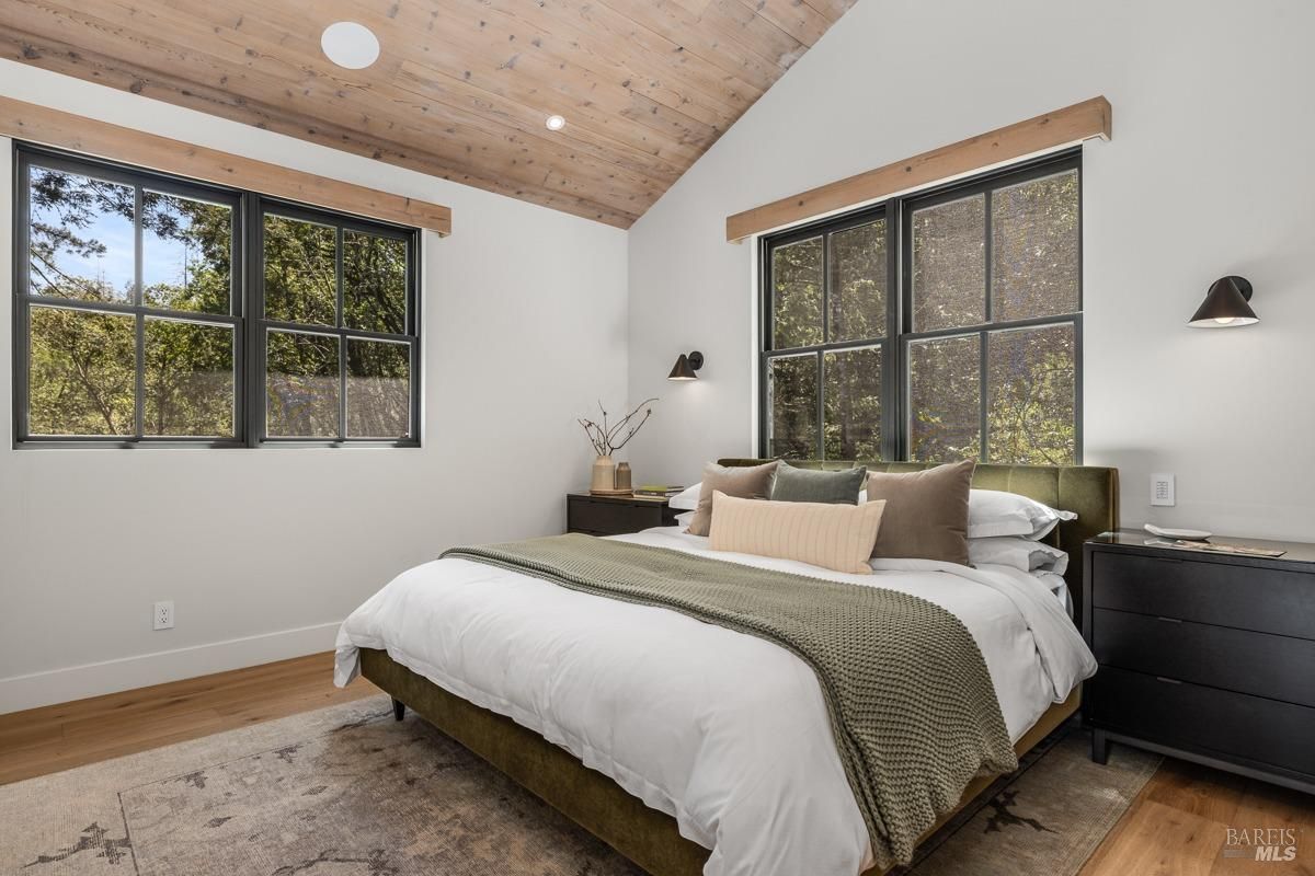  A calming bedroom featuring a vaulted wood-paneled ceiling, large windows, and neutral-toned furnishings. The design emphasizes simplicity and natural textures. 