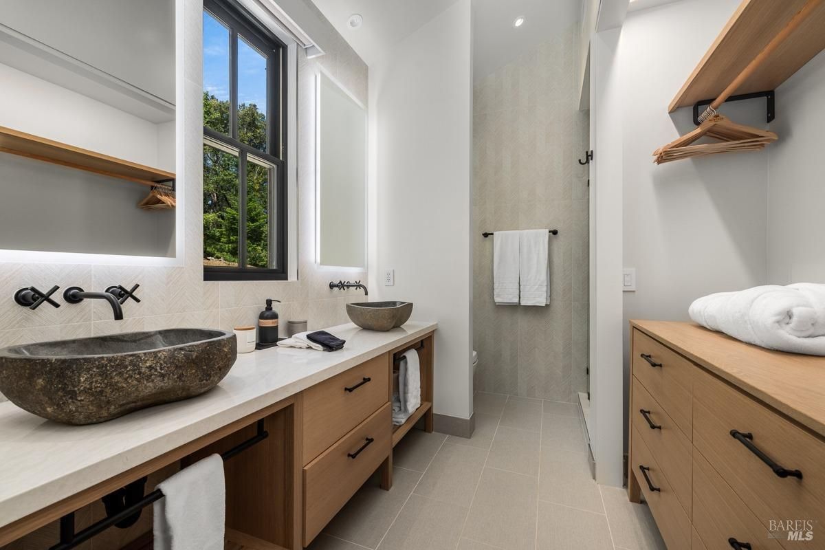  A contemporary bathroom featuring unique stone vessel sinks, black fixtures, and a warm wood vanity. The design integrates natural textures and materials, with a large window offering views of the greenery outside.