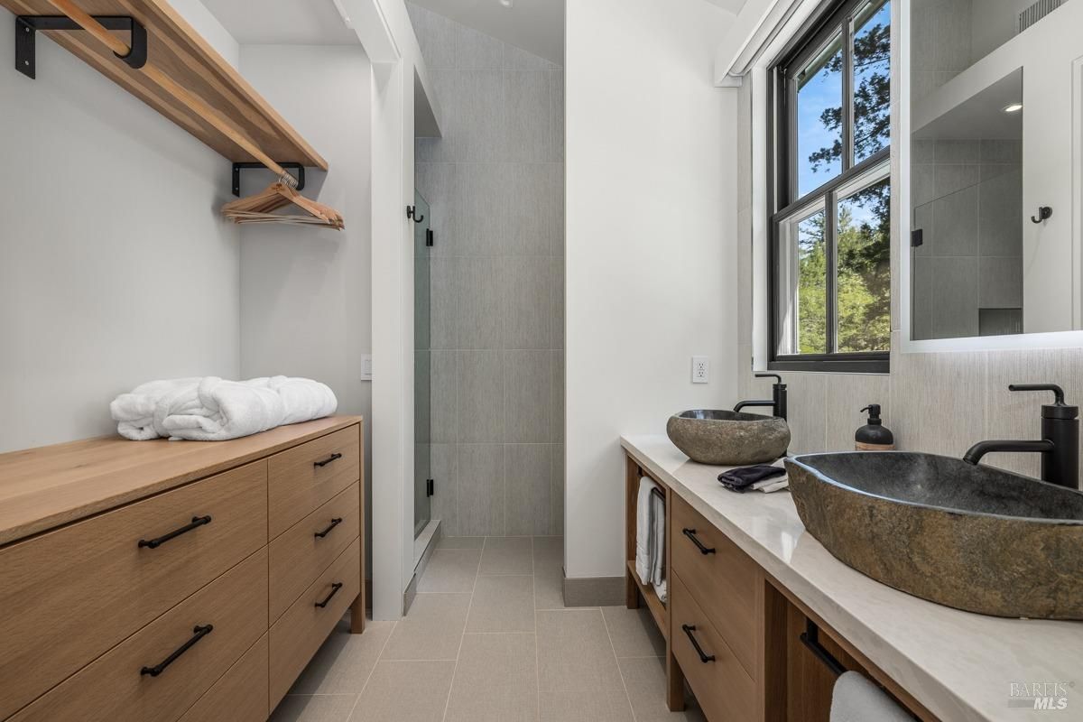 This minimalist bathroom includes wooden cabinetry, open shelving, and a walk-in shower. The stone sinks and black hardware add a rustic yet modern feel, complemented by a neutral tile floor.