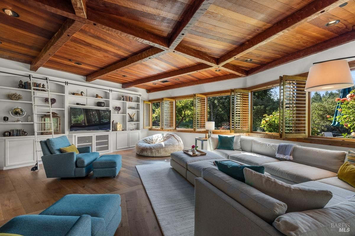 A spacious and inviting living room with a large sectional sofa, a bean bag chair, and a variety of armchairs and ottomans. The room features a wood-beamed ceiling, large windows with shutters, and built-in bookshelves. The overall atmosphere is cozy and inviting, perfect for relaxing and entertaining guests.