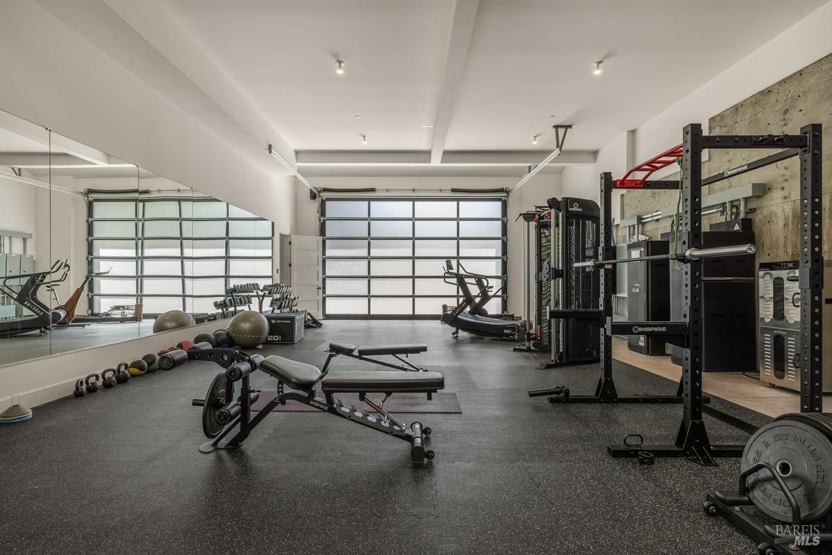 A spacious gym with rubber flooring, large mirrors, and industrial-style garage doors. The equipment is neatly organized, and the space has a clean, functional vibe with plenty of natural light. 