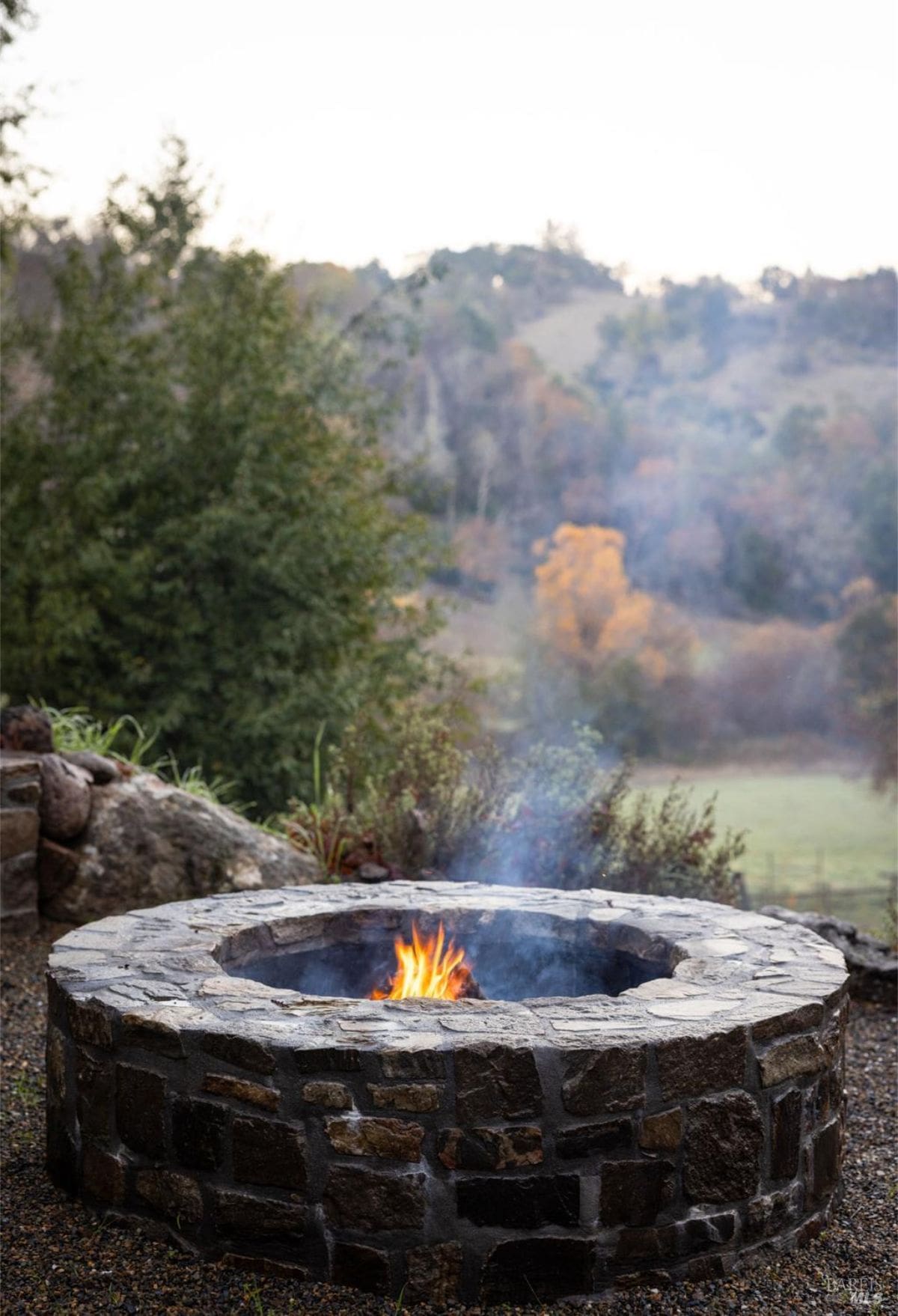 A stone fire pit in a picturesque outdoor setting, with views of rolling hills and autumn foliage. The fire pit is perfect for cozy evenings while enjoying nature.