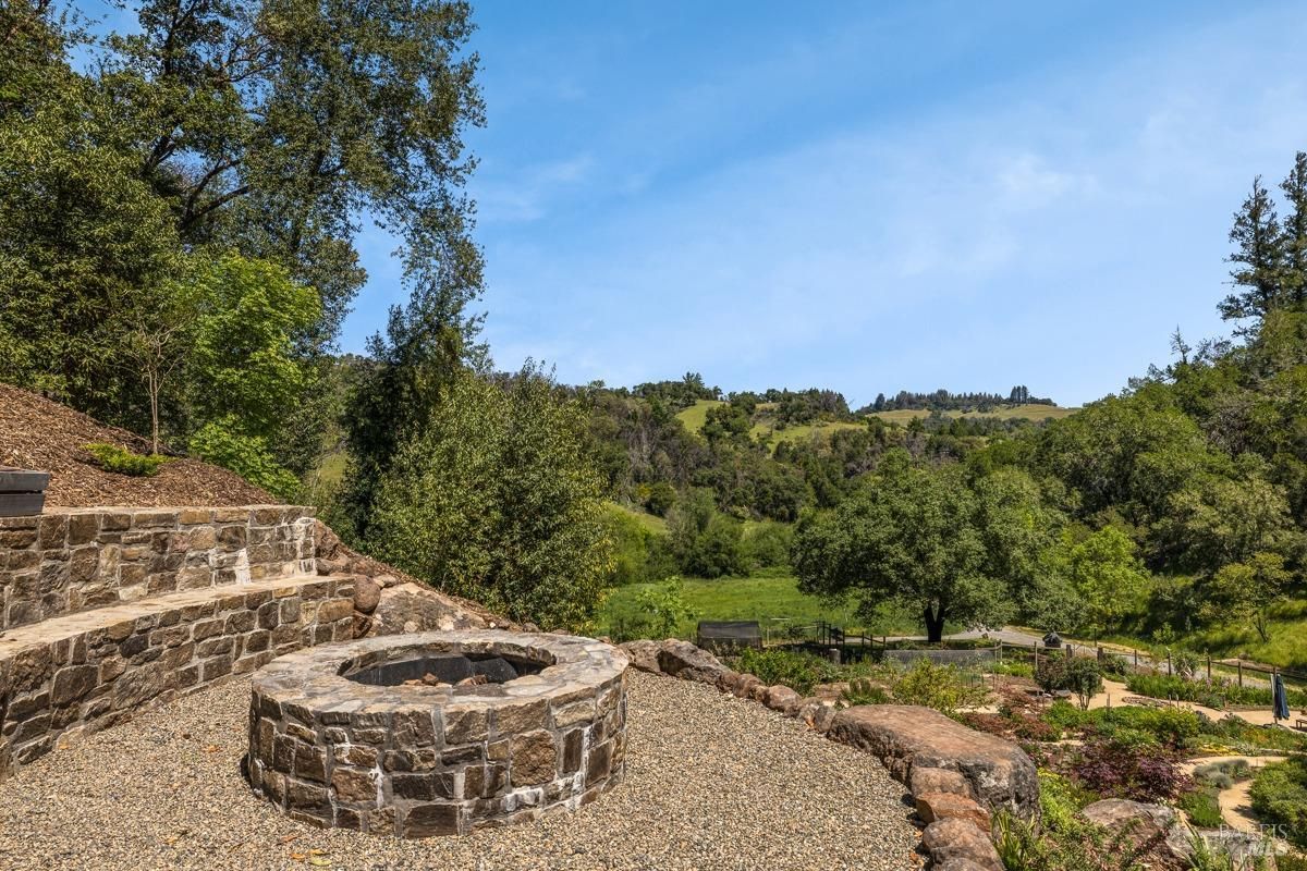 A fire pit made of stone surrounded by a gravel patio, set in a natural, outdoor environment. The backdrop includes rolling green hills and trees, emphasizing a peaceful and rural setting.