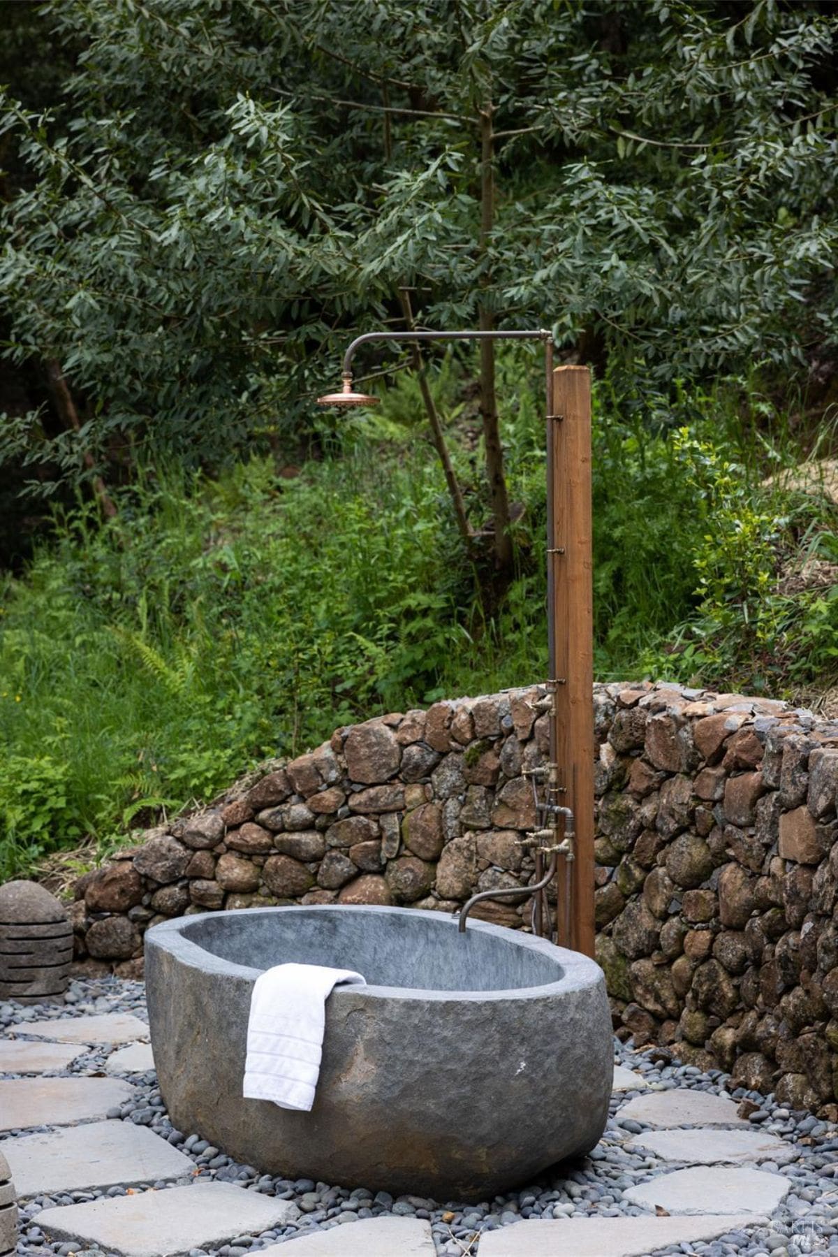 A rustic and serene outdoor bathing area featuring a stone tub surrounded by a stone wall and lush greenery. A wooden shower fixture adds a natural, spa-like vibe to the private setting.