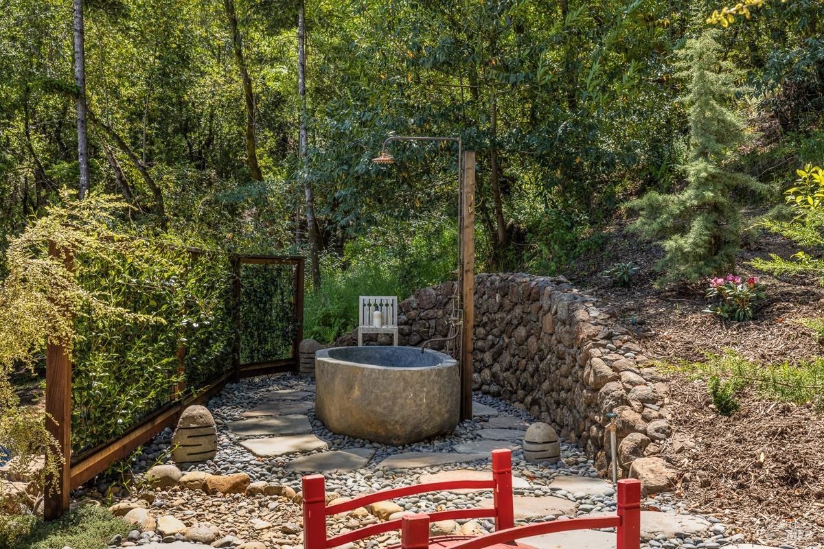  An extended view of the outdoor bath area, with red fencing and landscaping enhancing the serene environment. The stone tub and shower are nestled into a private, tranquil corner surrounded by plants.