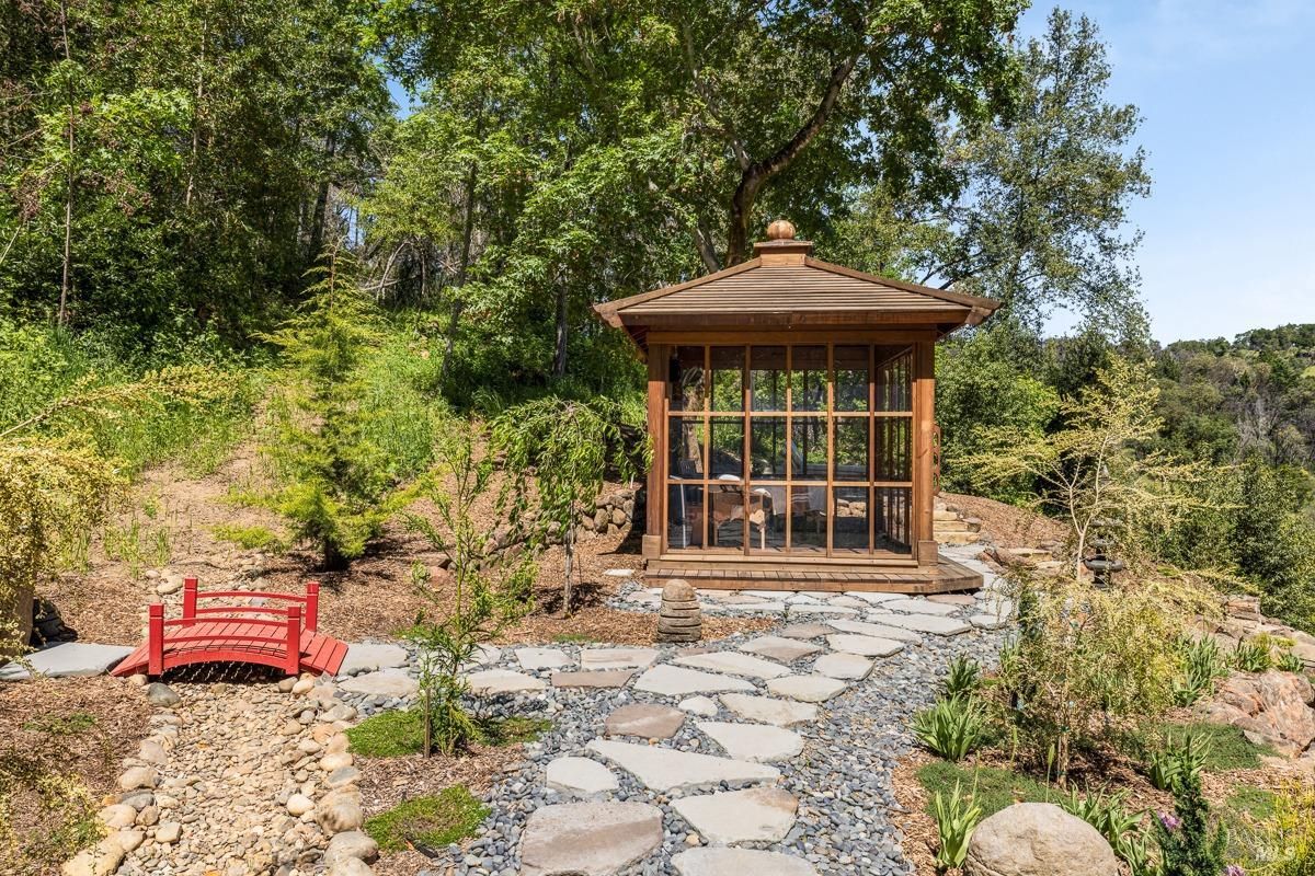 A wooden gazebo with glass panels, surrounded by a tranquil landscape featuring stone paths, trees, and a red footbridge. The setting evokes a peaceful and meditative atmosphere. 