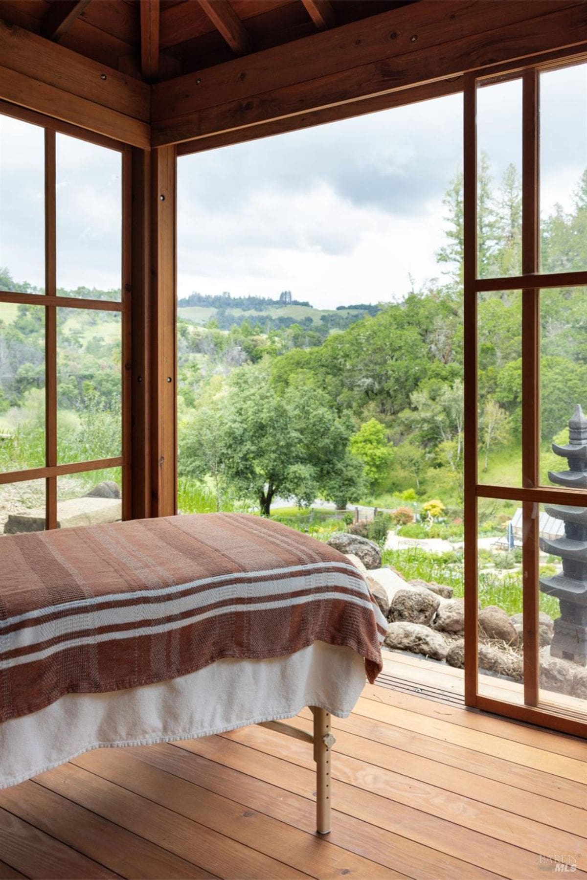 A serene outdoor space with a massage or spa bed overlooking lush greenery and rolling hills. The wooden-framed structure provides a rustic ambiance, while large windows bring in an abundance of natural light and emphasize the scenic environment.