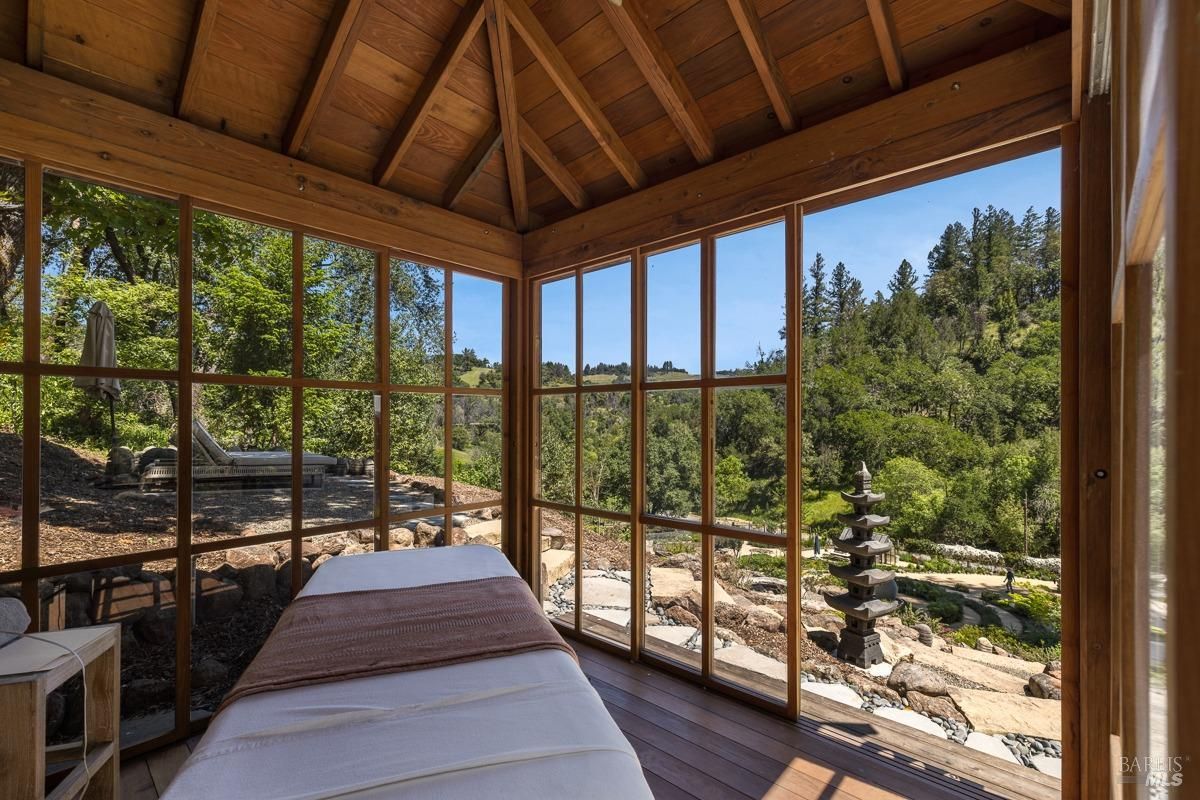 The interior of the wooden gazebo, featuring a massage table or resting area. The surrounding windows offer panoramic views of lush greenery and hills, creating a relaxing environment.