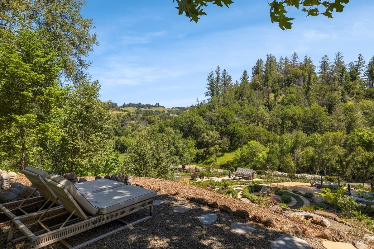 A scenic view of a relaxing spot featuring two lounge chairs overlooking a valley with dense greenery and rolling hills. The area seems designed for enjoying the peaceful surroundings and panoramic views of nature.