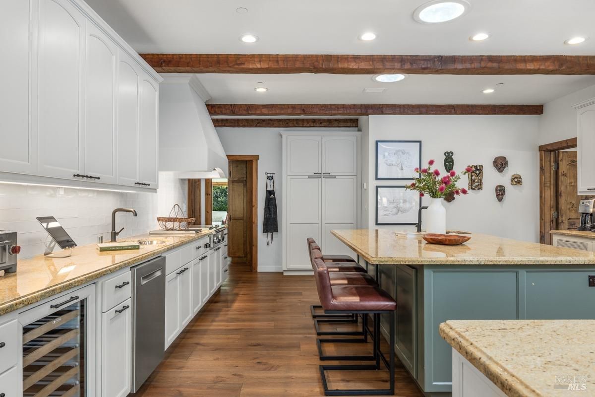  A bright kitchen with white cabinets, wooden beams on the ceiling, and a spacious island with seating. The countertops are beige granite, and the overall ambiance blends farmhouse charm with modern convenience.