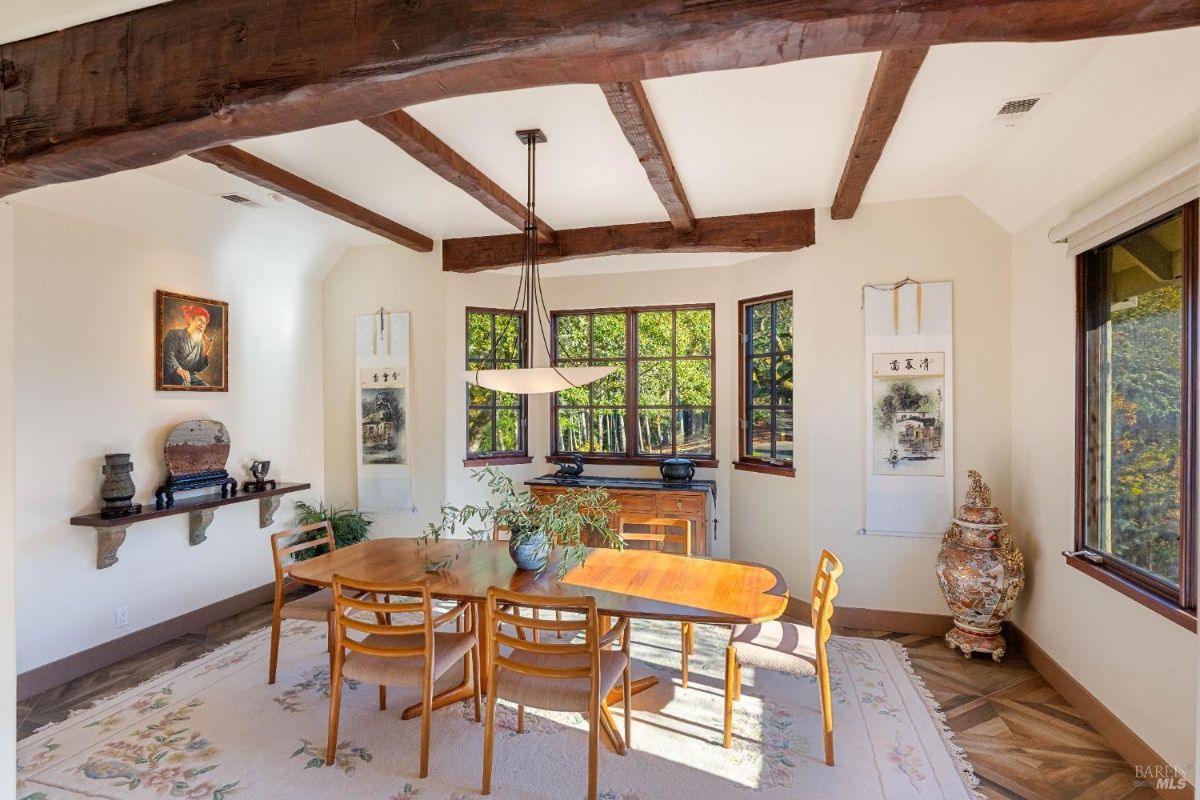 Dining room with exposed wooden beams, large windows, and a rectangular wooden table surrounded by chairs.