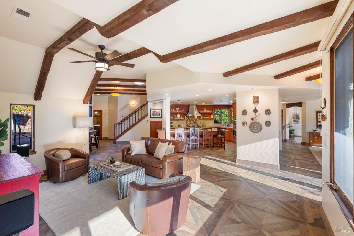 Open-concept room showing a living area, kitchen, and staircase with wooden beams on the ceiling.